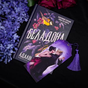 May include: A book titled "Беладонна" with a floral and bird design, next to a purple bookmark with a couple dancing. The bookmark has a purple tassel. Lilac flowers and dark red roses are in the background.