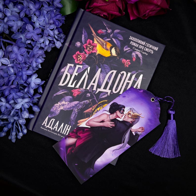 May include: A book titled "Беладонна" with a floral and bird design, next to a purple bookmark with a couple dancing. The bookmark has a purple tassel. Lilac flowers and dark red roses are in the background.