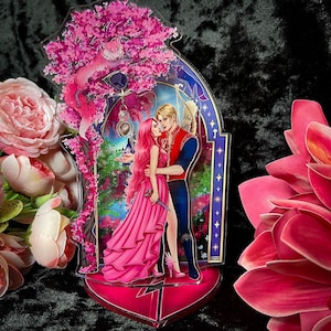 Könnte beinhalten: Ein rosa-blauer Acryl-Ständer mit einem Paar, das sich vor einem rosa Kirschblütenbaum umarmt. Das Paar hält einen Dolch und einen Bogen und Pfeil. Der Ständer hat eine herzförmige Basis.