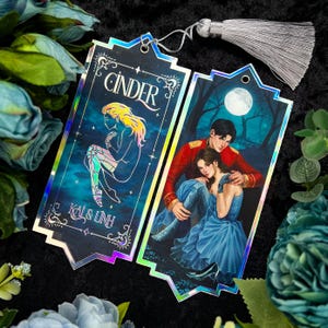 Könnte beinhalten: Zwei schimmernde Lesezeichen mit Figuren aus dem Buch "Cinder". Das erste Lesezeichen zeigt eine junge Frau mit einem mechanischen Bein, mit dem Text "Cinder" und "Kai & Linh". Das zweite Lesezeichen zeigt ein Paar in einer Waldkulisse mit einem Vollmond im Hintergrund.