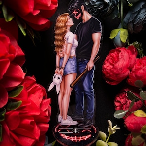 May include: A decorative acrylic figure featuring a couple embracing, with a red X mask and a bunny mask. The man holds a baseball bat. The base is heart-shaped with a red glowing smile design. Surrounded by red and blue flowers.