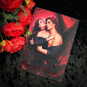 May include: A print of a couple embracing, the woman with long dark hair and the man with dark hair and wings. The background is a red and orange gradient with black feathers. The text "Oragya & Rahln" is in the bottom corner.