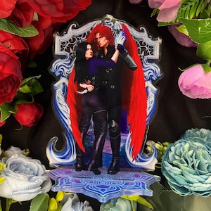 Könnte beinhalten: Acrylständer mit einem Paar in einer romantischen Umarmung, wobei die männliche Figur große rote Flügel trägt. Das Kunstwerk hat ein Fantasy-Thema, wobei die Figuren dunkle Kleidung tragen. Der Ständer hat einen dekorativen Sockel und Rahmen mit blauen Akzenten.