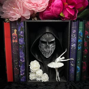 May include: A black box display featuring a hooded mask with a detailed face design, a white ballerina figurine, and white roses. The display is flanked by decorative books and topped with pink and magenta flowers.