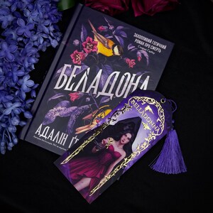 May include: A book and bookmark with the title "Belladonna". The book cover is dark with floral and bird illustrations. The bookmark features a woman in a red dress. The text on the book is in a foreign language.
