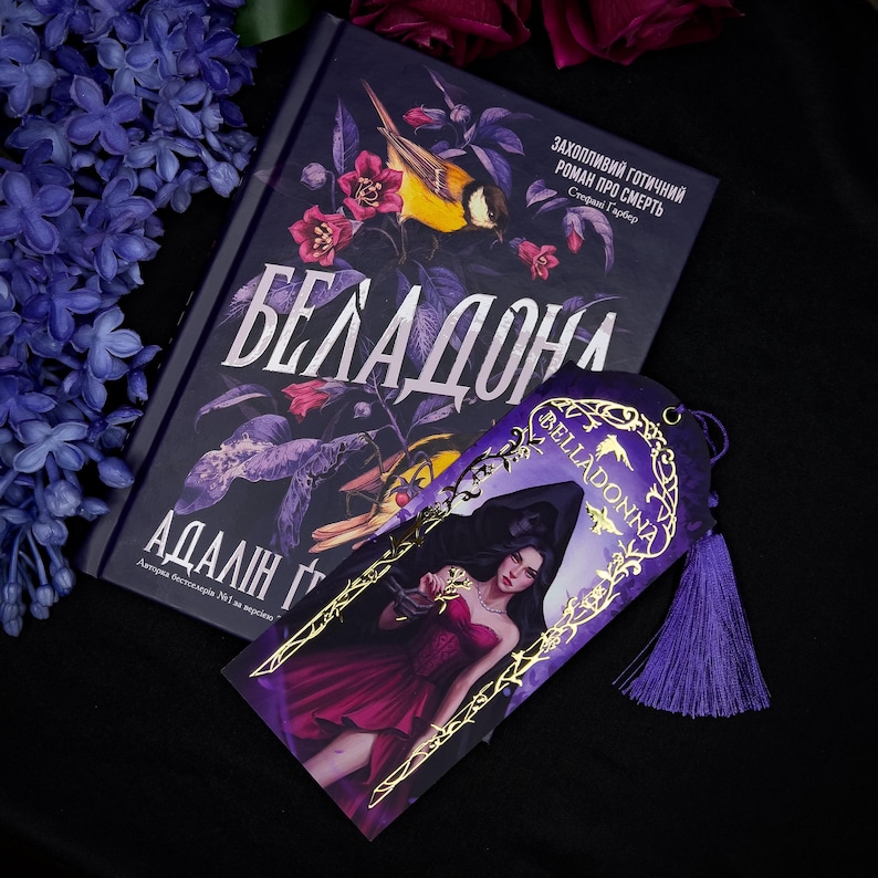 May include: A book and bookmark with the title "Belladonna". The book cover is dark with floral and bird illustrations. The bookmark features a woman in a red dress. The text on the book is in a foreign language.