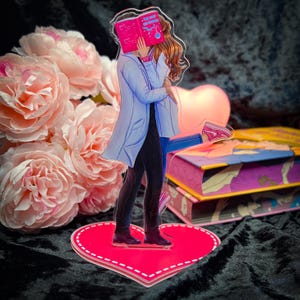 Puede incluir: Una base rosa en forma de corazón con un borde blanco punteado sostiene a una pareja que sostiene un libro. El hombre lleva una bata de laboratorio blanca y pantalones negros. La mujer lleva una camisa azul y pantalones negros. El libro es rosa con una cubierta azul y el texto "The Book of Enchantment" en la cubierta.