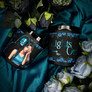 Op de afbeelding: Twee zwarte kaarsglazen met bloemen- en grafische ontwerpen. Eén pot heeft een vrouw en een zeisfiguur. De andere pot heeft de woorden "Lights Out" in teal. Beide potten zijn omgeven door blauwe rozen.