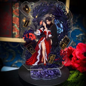 May include: A clear acrylic standee featuring a digital illustration of a person in a red dress and a person in a black outfit embraced by a dark, shadowy wolf-like creature. Four tarot cards with gold symbols float around them against a dark background.