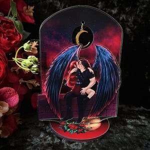 Puede incluir: Una pareja con alas se besa bajo una luna creciente. La pareja está silueteada contra un cielo oscuro y estrellado. La imagen está impresa en un soporte de acrílico transparente.