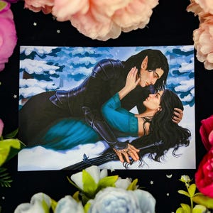 Può includere: Una stampa artistica raffigurante una scena romantica con due figure in un paesaggio innevato. Una figura, in armatura nera, abbraccia l'altra, che indossa un abito turchese. L'opera presenta lunghi capelli neri e un tema fantasy.