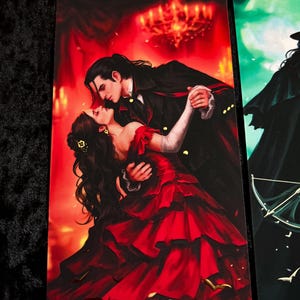 May include: A bookmark featuring a romantic illustration of a couple dancing in a ballroom. The woman wears a red gown, and the man is in a black coat. The word "Dracula" is at the bottom. A gold-colored eyelet is at the top.