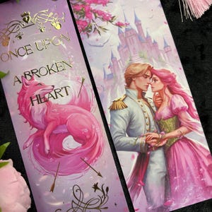 Peut inclure: Deux marque-pages d&eacute;coratifs. L'un pr&eacute;sente une illustration de renard rose avec le texte "ONCE UPON A BROKEN HEART" en lettres dor&eacute;es. L'autre repr&eacute;sente un couple devant un ch&acirc;teau, avec des fleurs roses. Les marque-pages ont un th&egrave;me fantastique.