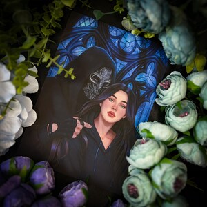 May include: A woman with long dark hair is wearing a black hooded jacket and looking at the camera. She is standing next to a person wearing a black hooded cloak with a silver mask. The background is a dark blue stained glass window.