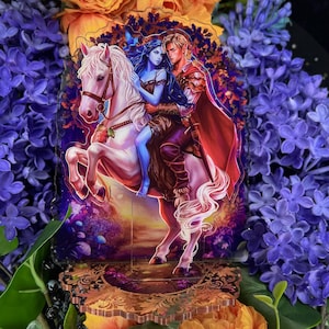 Puede incluir: Un soporte acrílico de temática fantástica que representa a una pareja montada a caballo blanco. La mujer tiene la piel y el pelo azules, mientras que el hombre lleva una armadura roja y una capa. El soporte tiene una base ornamentada y está rodeado de flores moradas.