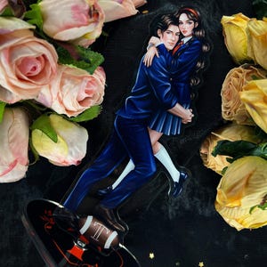 May include: An acrylic stand featuring a couple embracing, dressed in school uniforms. The man wears a navy blue suit, and the woman wears a blue skirt and blazer. The base is black with a football design and the letter "T".