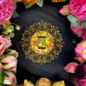 Pu&ograve; includere: Un sottobicchiere in acrilico trasparente con un design a mandala dorato. Il centro presenta un motivo floreale nei toni del giallo, arancione e verde. Il sottobicchiere ha due fessure ed &egrave; circondato da fiori colorati su sfondo nero.