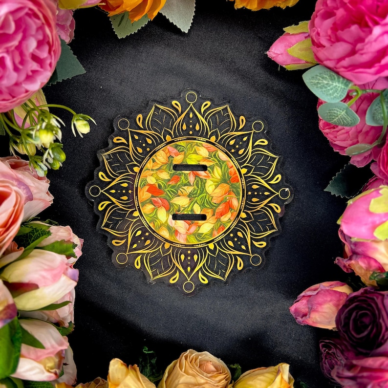 Pu&ograve; includere: Un sottobicchiere in acrilico trasparente con un design a mandala dorato. Il centro presenta un motivo floreale nei toni del giallo, arancione e verde. Il sottobicchiere ha due fessure ed &egrave; circondato da fiori colorati su sfondo nero.