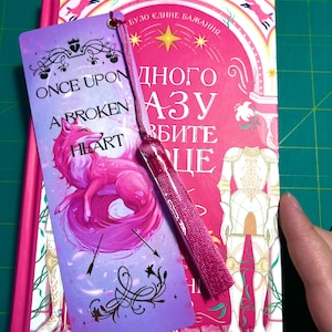 Peut inclure: Un marque-page rose et violet avec un pompon est pos&eacute; sur un livre. Le marque-page pr&eacute;sente une illustration de renard rose et le texte "ONCE UPON A BROKEN HEART". Le livre a une couverture rose et blanche avec du texte en langue &eacute;trang&egrave;re.