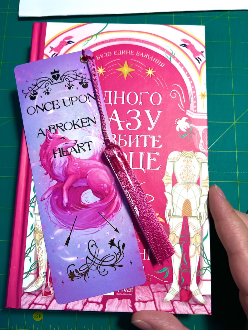 Peut inclure: Un marque-page rose et violet avec un pompon est pos&eacute; sur un livre. Le marque-page pr&eacute;sente une illustration de renard rose et le texte "ONCE UPON A BROKEN HEART". Le livre a une couverture rose et blanche avec du texte en langue &eacute;trang&egrave;re.