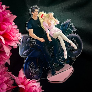 Puede incluir: Una pareja en una motocicleta negra. La mujer lleva un vestido blanco y una venda roja en los ojos. El hombre lleva una camisa negra y pantalones vaqueros. La motocicleta tiene una base rosa.