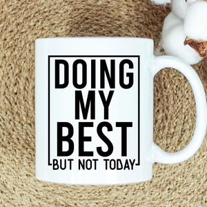 May include: White ceramic mug with the text "DOING MY BEST BUT NOT TODAY" in black, within a black rectangular border. The mug has a curved handle and is set against a neutral background.
