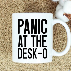 May include: White ceramic mug with the text "PANIC AT THE DESK-O" in bold black letters. The mug has a curved handle and is set against a neutral background with cotton accents.
