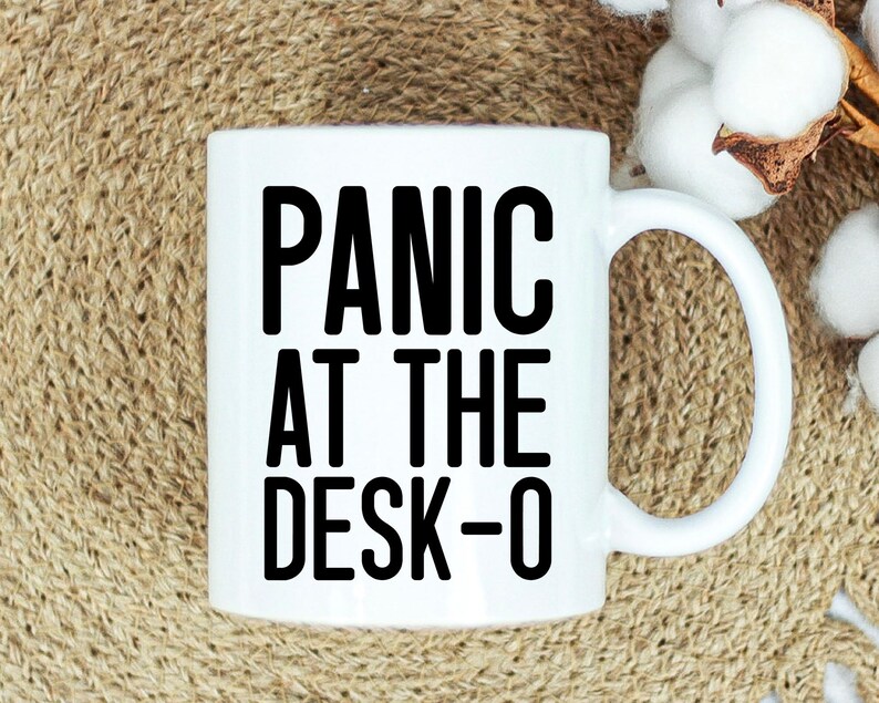 May include: White ceramic mug with the text "PANIC AT THE DESK-O" in bold black letters. The mug has a curved handle and is set against a neutral background with cotton accents.