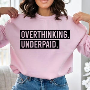May include: A light pink sweatshirt with the text "OVERTHINKING. UNDERPAID." in a black, bold font. The sweatshirt has long sleeves and a crew neck. The model is wearing blue jeans.