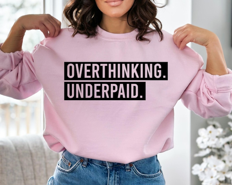 May include: A light pink sweatshirt with the text "OVERTHINKING. UNDERPAID." in a black, bold font. The sweatshirt has long sleeves and a crew neck. The model is wearing blue jeans.