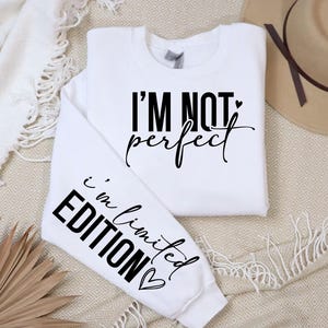 Könnte beinhalten: Weißes Sweatshirt mit dem schwarzen Text "I'M NOT perfect" auf der Vorderseite. Der linke Ärmel hat den schwarzen Text "i'm limited EDITION". Ein beiger Hut mit braunem Band ist im Hintergrund.
