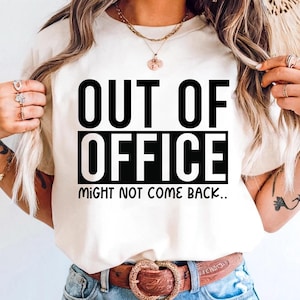 May include: Cream-colored t-shirt with the text "OUT OF OFFICE" in bold black letters, stacked over "MIGHT NOT COME BACK." The shirt is worn with blue jeans and a brown belt with a decorative buckle. The overall theme is casual wear.