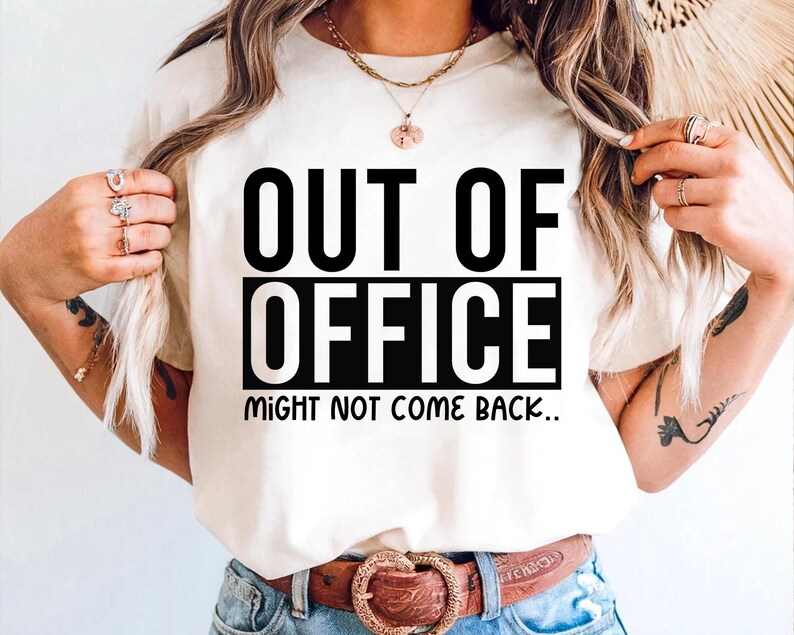 May include: Cream-colored t-shirt with the text "OUT OF OFFICE" in bold black letters, stacked over "MIGHT NOT COME BACK." The shirt is worn with blue jeans and a brown belt with a decorative buckle. The overall theme is casual wear.