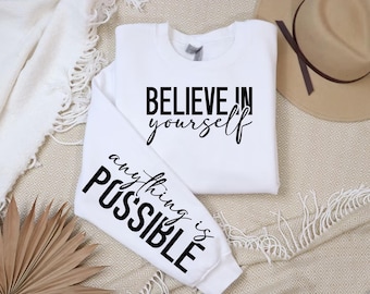 May include: White sweatshirt with the words "BELIEVE in yourself" printed in black script on the front. The left sleeve has the words "anything is POSSIBLE" printed in a bold, black font. A tan hat is visible in the background.