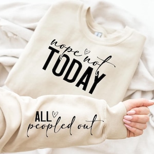 以下が含まれることがあります： ベージュ色のスウェットシャツ。黒い筆記体で「nope not TODAY」と「ALL peopled out」の文字がプリントされています。袖が見えており、手とマニキュアされた爪があります。スウェットシャツは白い表面に折りたたまれています。