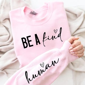 May include: A light pink sweatshirt with the words "BE A kind human" printed in black script. The word "kind" has a heart above the "i". The sleeve also has the word "human" with a heart above the "u".