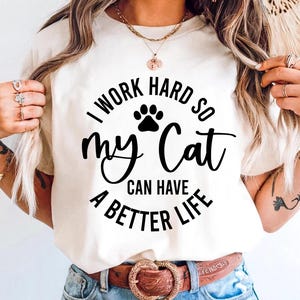 May include: Cream-colored t-shirt with the text "I WORK HARD SO my Cat CAN HAVE A BETTER LIFE" in black. The design includes a paw print. The shirt has a round neckline and short sleeves.