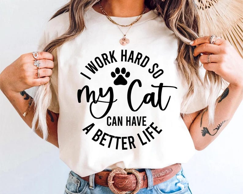 May include: Cream-colored t-shirt with the text "I WORK HARD SO my Cat CAN HAVE A BETTER LIFE" in black. The design includes a paw print. The shirt has a round neckline and short sleeves.