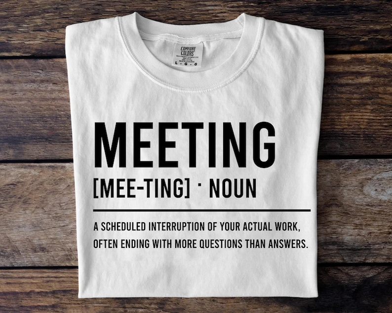 May include: White t-shirt with the word "MEETING" in large black letters. Below the word is the phonetic pronunciation and the word "NOUN". The shirt's definition of a meeting is printed below the word "NOUN".