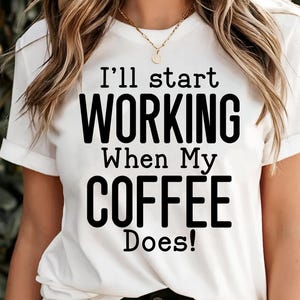 May include: White t-shirt with the text "I'll start WORKING When My COFFEE Does!" in black. The shirt has short sleeves and a round neckline. A gold necklace is visible.