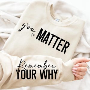 以下が含まれることがあります： 「you matter」と「Remember YOUR WHY」の文字が黒字でプリントされたベージュ色のスウェットシャツ。スウェットシャツは折りたたまれており、袖とテキストが見える。全体的なテーマはモチベーション。