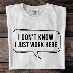 May include: White t-shirt with the text "I DON'T KNOW I JUST WORK HERE" in a black speech bubble. The shirt is folded and lies on a wooden surface. The shirt has a crew neck and a tag.