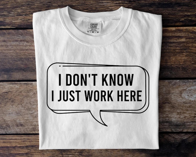 May include: White t-shirt with the text "I DON'T KNOW I JUST WORK HERE" in a black speech bubble. The shirt is folded and lies on a wooden surface. The shirt has a crew neck and a tag.