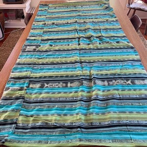 May include: A rectangular textile featuring a horizontal striped pattern in shades of turquoise, lime green, black, and white. The fabric displays repeating geometric and abstract motifs integrated into the bands of color across the entire surface.