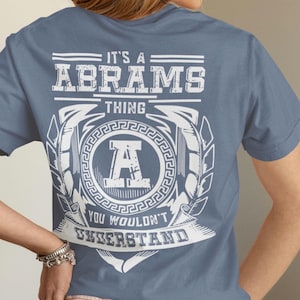 May include: A blue t-shirt with a white graphic design on the back. The design includes the text "IT'S A ABRAMS THING YOU WOULDN'T UNDERSTAND" with a large letter "A" in the center, surrounded by a laurel wreath.