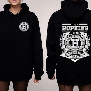 Könnte beinhalten: Schwarzer Kapuzenpullover mit einem weißen, kreisförmigen Emblem auf der Vorderseite und einem größeren Design auf der Rückseite. Das Rückendesign enthält den Text "IT'S A HOPKINS THING YOU WOULDN'T UNDERSTAND" in dekorativer Schrift.