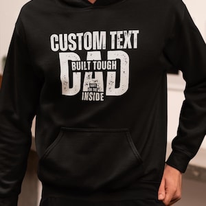 May include: Black hoodie with a front pocket and white text that reads "CUSTOM TEXT" above "BUILT TOUGH DAD SOFT ON THE INSIDE". The hoodie is a casual garment.