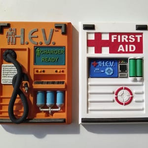Peut inclure: Deux plaques murales imprimées en 3D, l'une orange et l'autre blanche. La plaque orange porte le texte "H.E.V." et "CHARGER READY". La plaque blanche a une croix rouge et le texte "FIRST AID". Les deux plaques ont un petit rectangle bleu avec le texte "H.E.V." dessus.