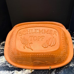 May include: An orange-brown ceramic Schlemmer Topf, a lidded cooking pot. The lid features embossed text and illustrations of corn, a garlic bulb, and a pumpkin. The pot has a rectangular shape with rounded edges and a textured surface.