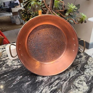 May include: A large, round copper pan with a hammered interior and brass handles. The pan is displayed on a black and white marble surface. The pan is a kitchenware item.